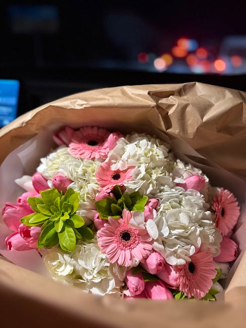A bouquet of fresh white hydrangeas and pink gerbera daisies, arranged with bright green foliage, wrapped in brown kraft paper with white tissue paper accents. The flowers are vibrant and lush, showca