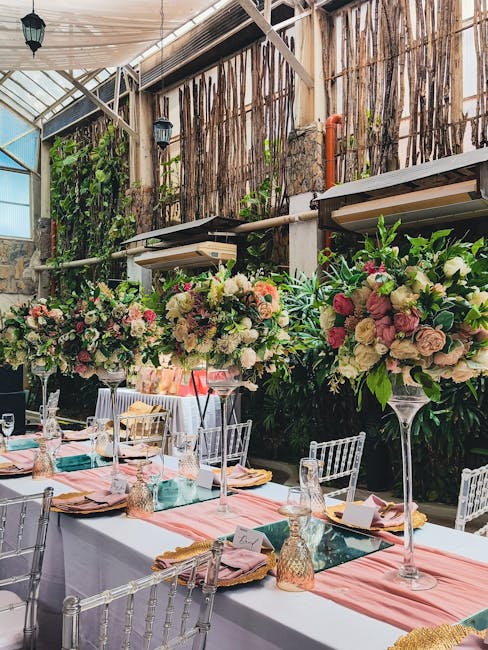 A floral arrangement featuring large, lush bouquets of pastel-colored roses, peonies, and hydrangeas with shades of pink, cream, and soft peach, arranged on tall glass vases as centerpieces. The flowe
