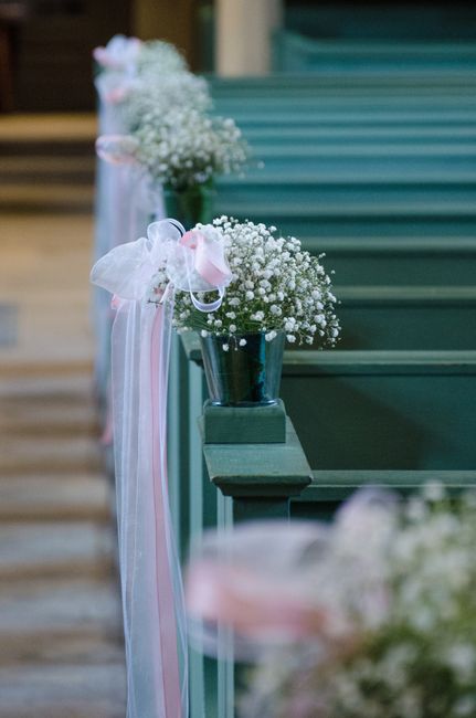 A series of small floral arrangements placed in dark metal vases, each adorned with delicate white baby's breath flowers and soft pink and white ribbon bows, lining a dark green wooden pew at a rivers